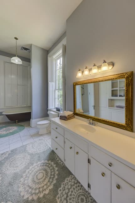 claw footed tub in back, large vanity with white cabinet doors, mirror with lots of lights, rug on floor
