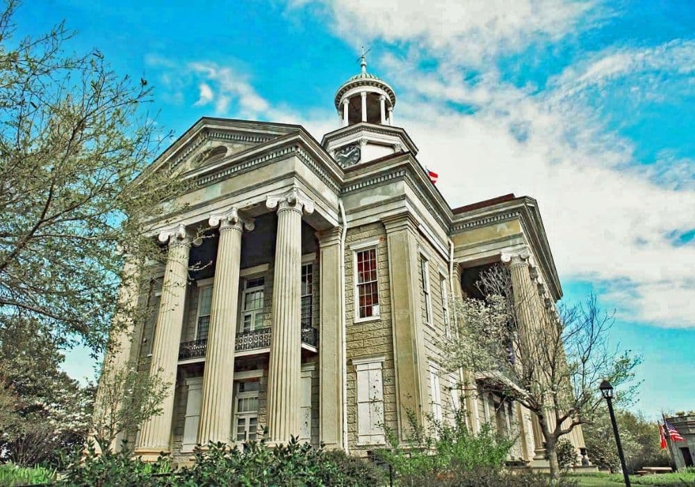 Historic building with tall columns and a clock tower, surrounded by greenery under a blue sky.