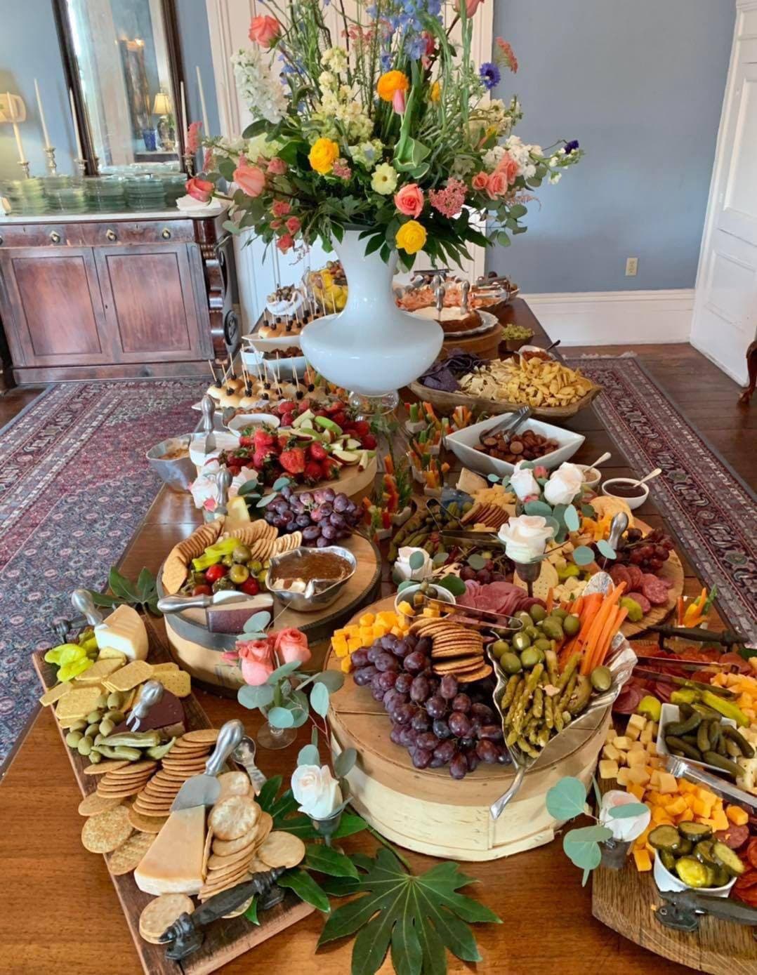 A lavish spread of assorted cheeses, meats, fruits, and vegetables, elegantly arranged on a wooden table with a large floral centerpiece.