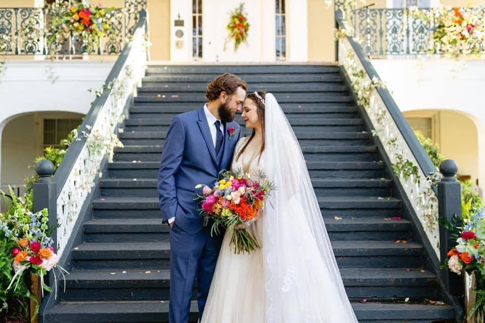 A bride and groom share a tender moment on the steps of a decorated venue, surrounded by floral arrangements.