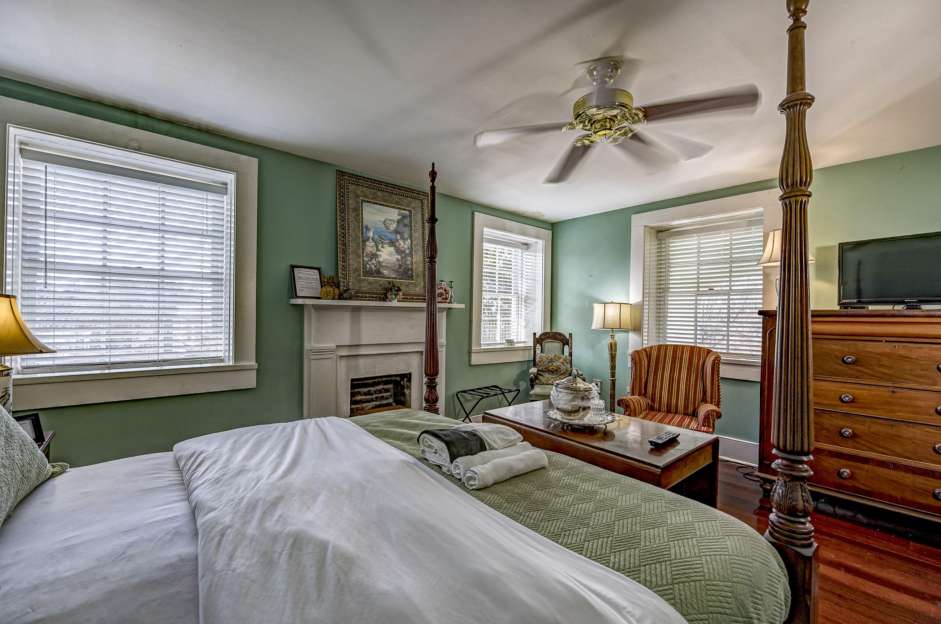 Cozy bedroom with a four-poster bed, green walls, and large windows, featuring a fireplace and comfortable seating.