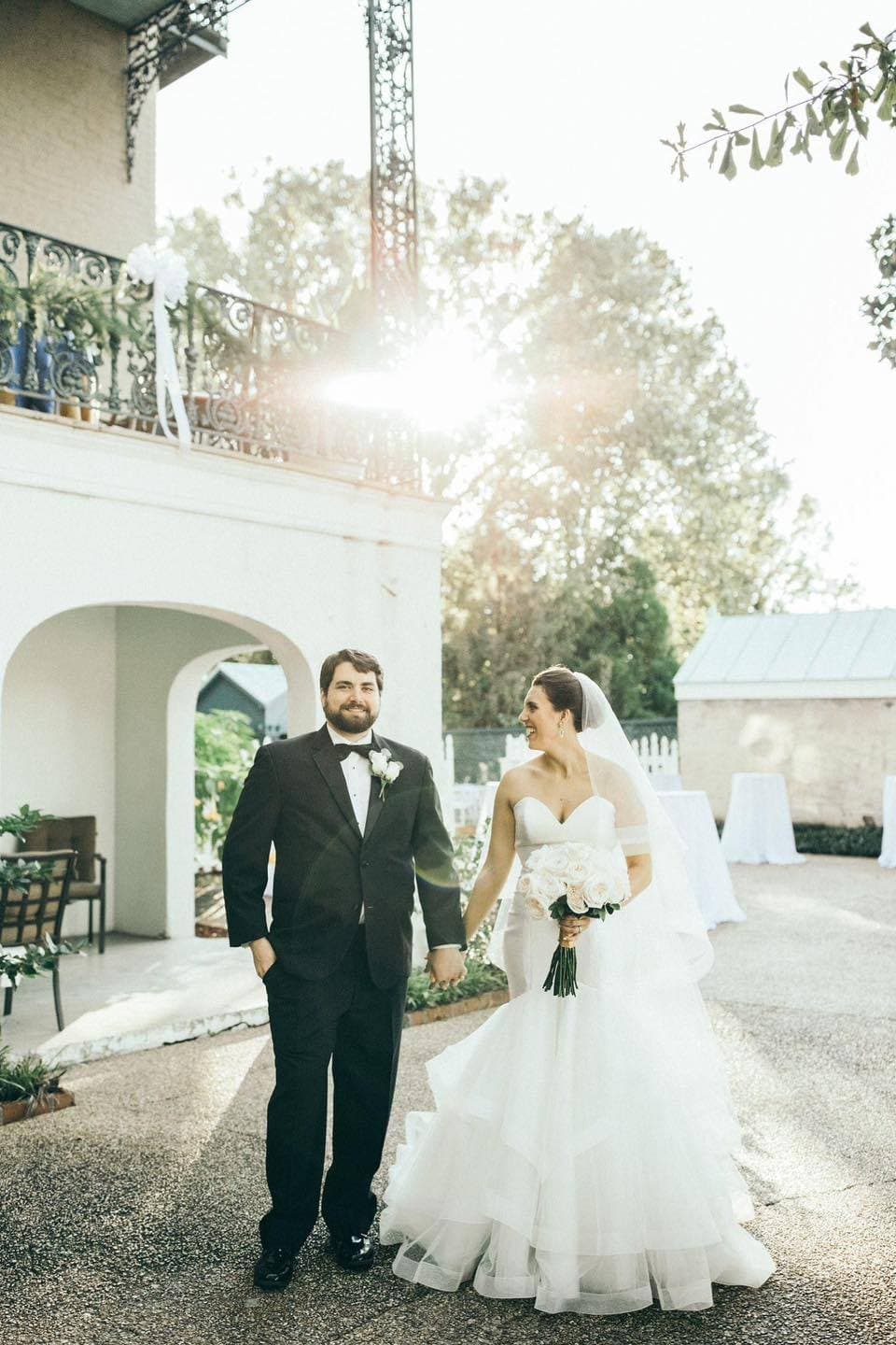 A couple in wedding attire holds hands, smiling at each other in a sunlit outdoor setting.