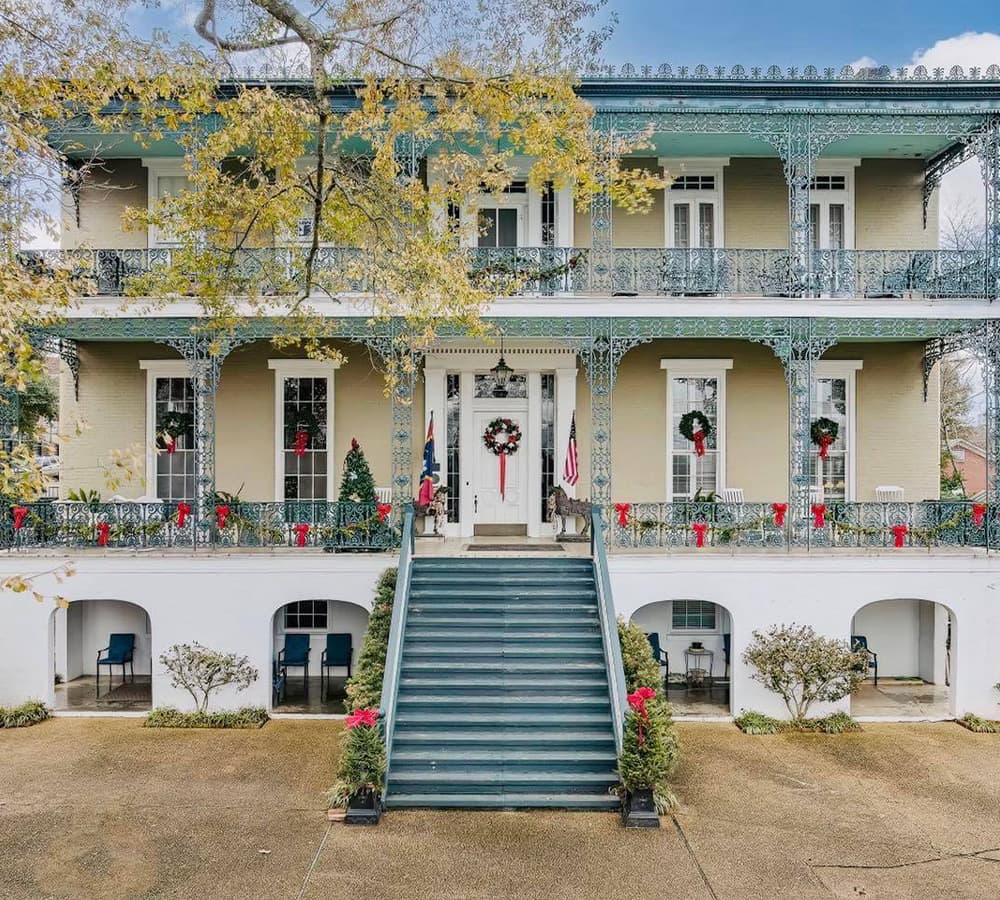 A large, decorative two-story house with a green balcony, festive wreaths, and a staircase leading to the entrance.