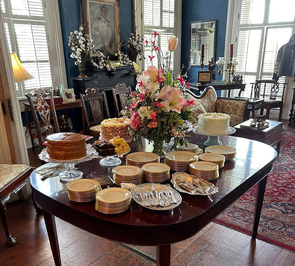 A beautifully arranged dining table with an assortment of cakes, elegant plates, and a vibrant floral centerpiece in a cozy living room.