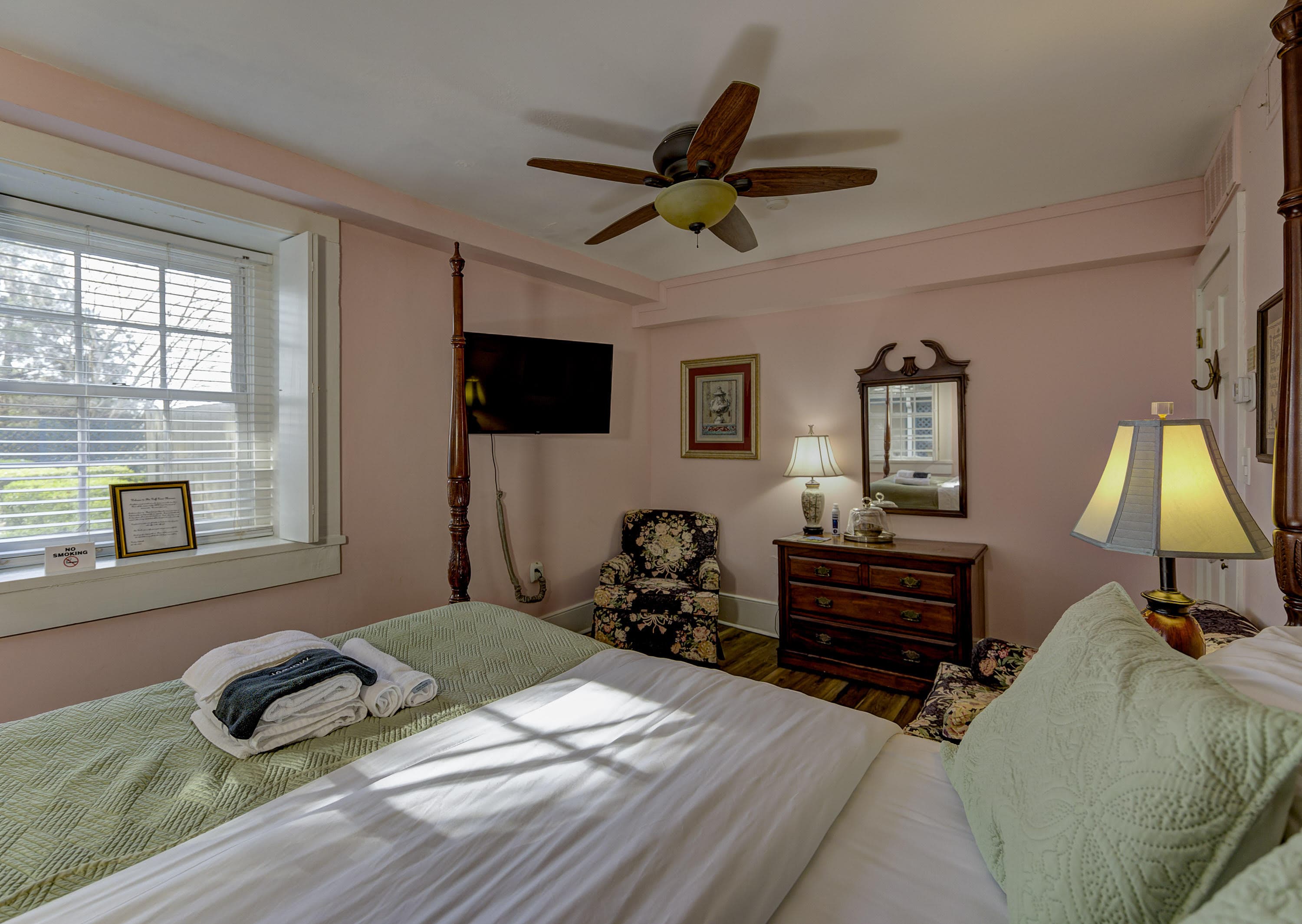 Cozy bedroom with a pink wall, a queen bed, vintage furniture, a ceiling fan, and natural light from a window.