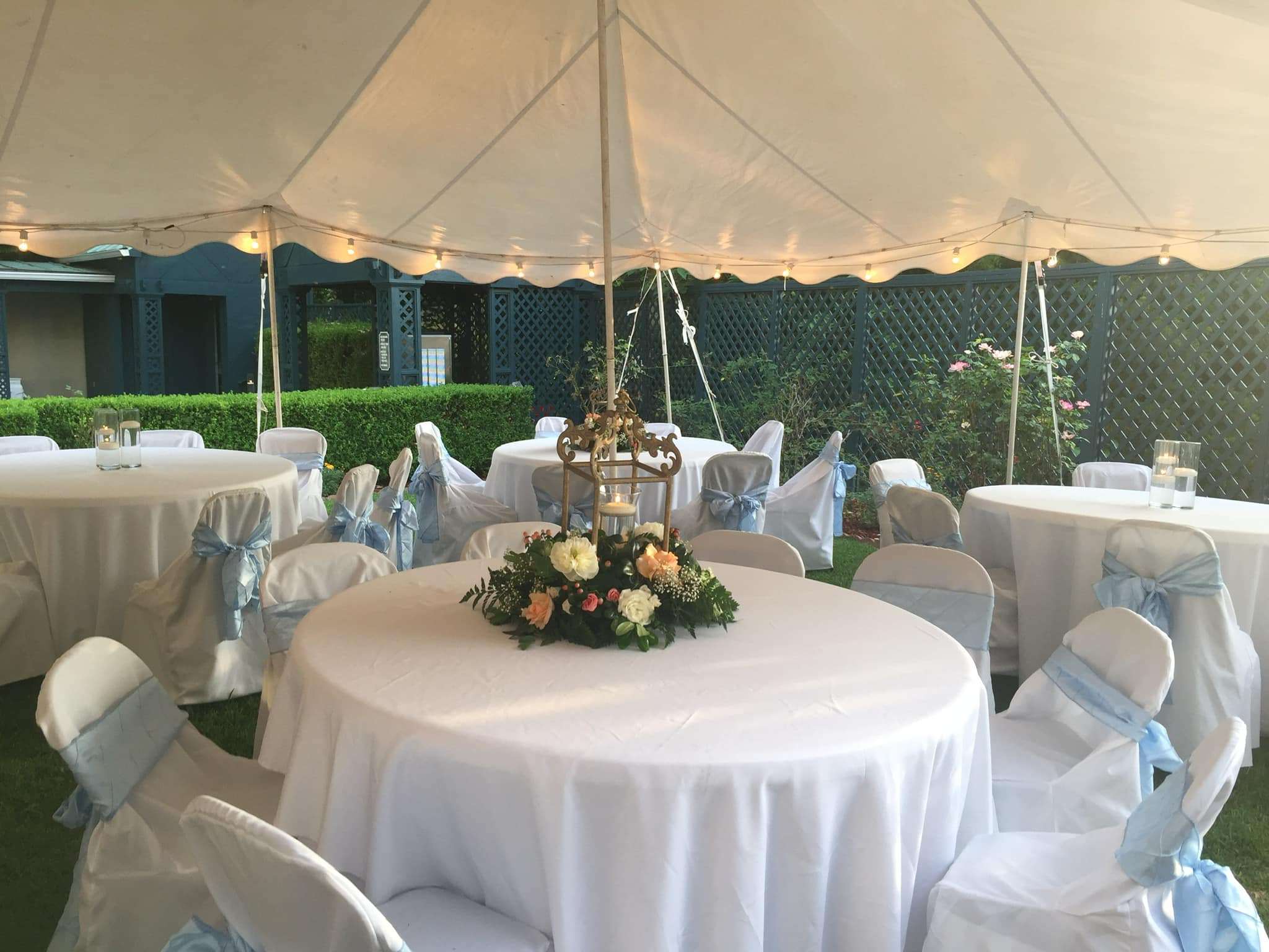 A decorated outdoor dining area under a tent, featuring round tables with white tablecloths and blue sashes.