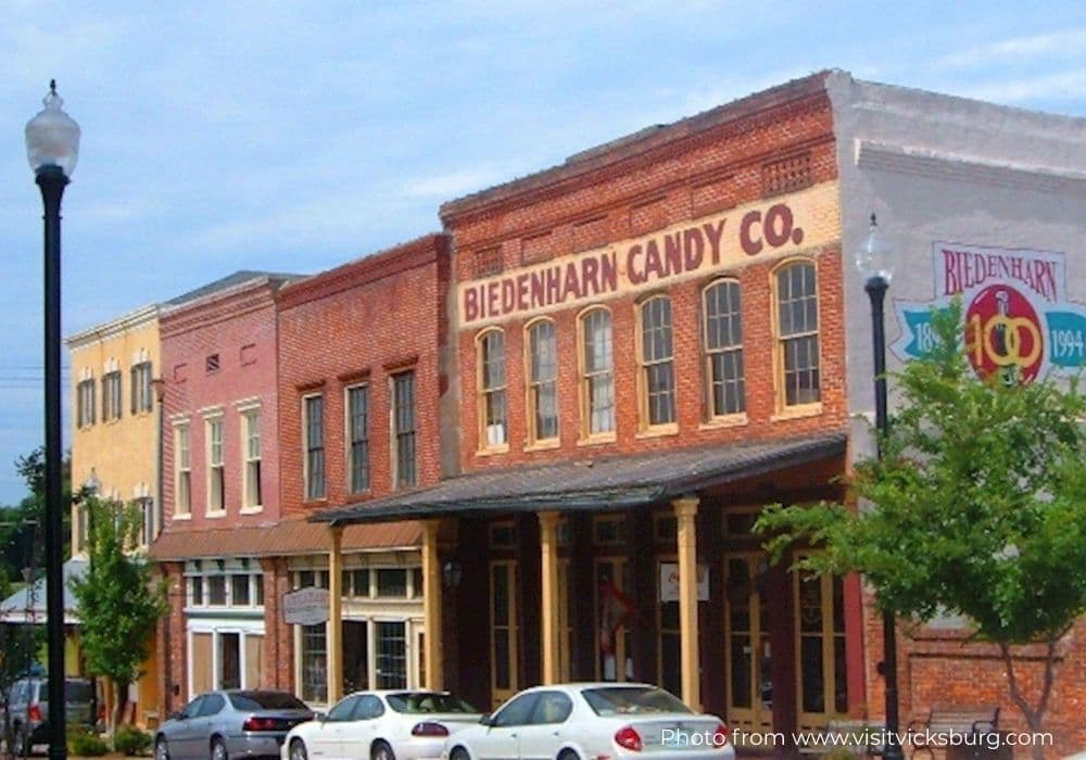 A brick building with the sign "Biedenharn Candy Co." and several storefronts, surrounded by trees and parked cars.