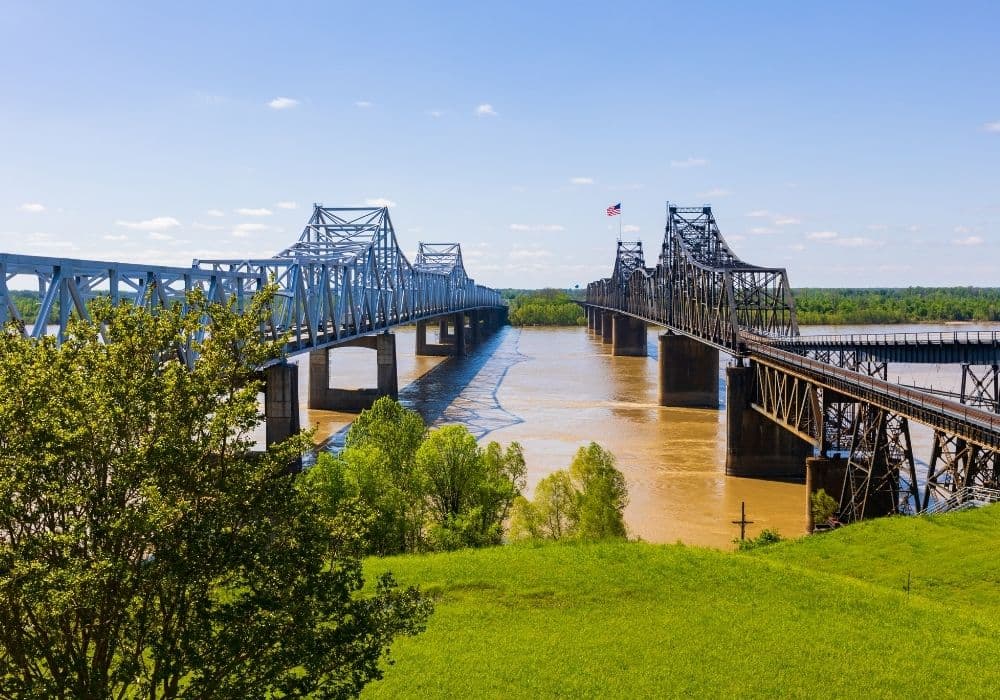 A scenic view of two bridges spanning a wide river, surrounded by greenery and a clear blue sky.