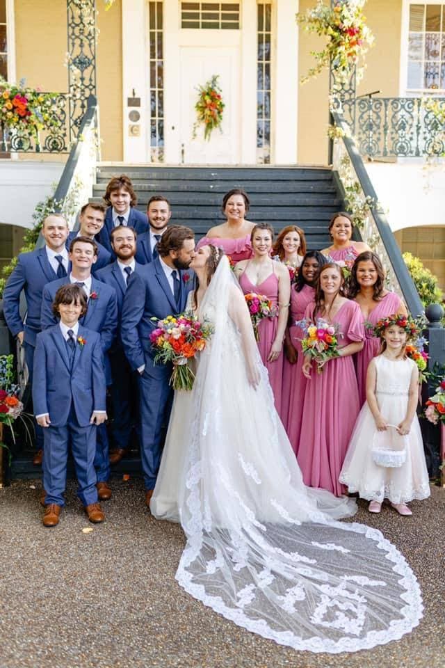 A bride and groom kiss in front of their wedding party on a staircase adorned with flowers.
