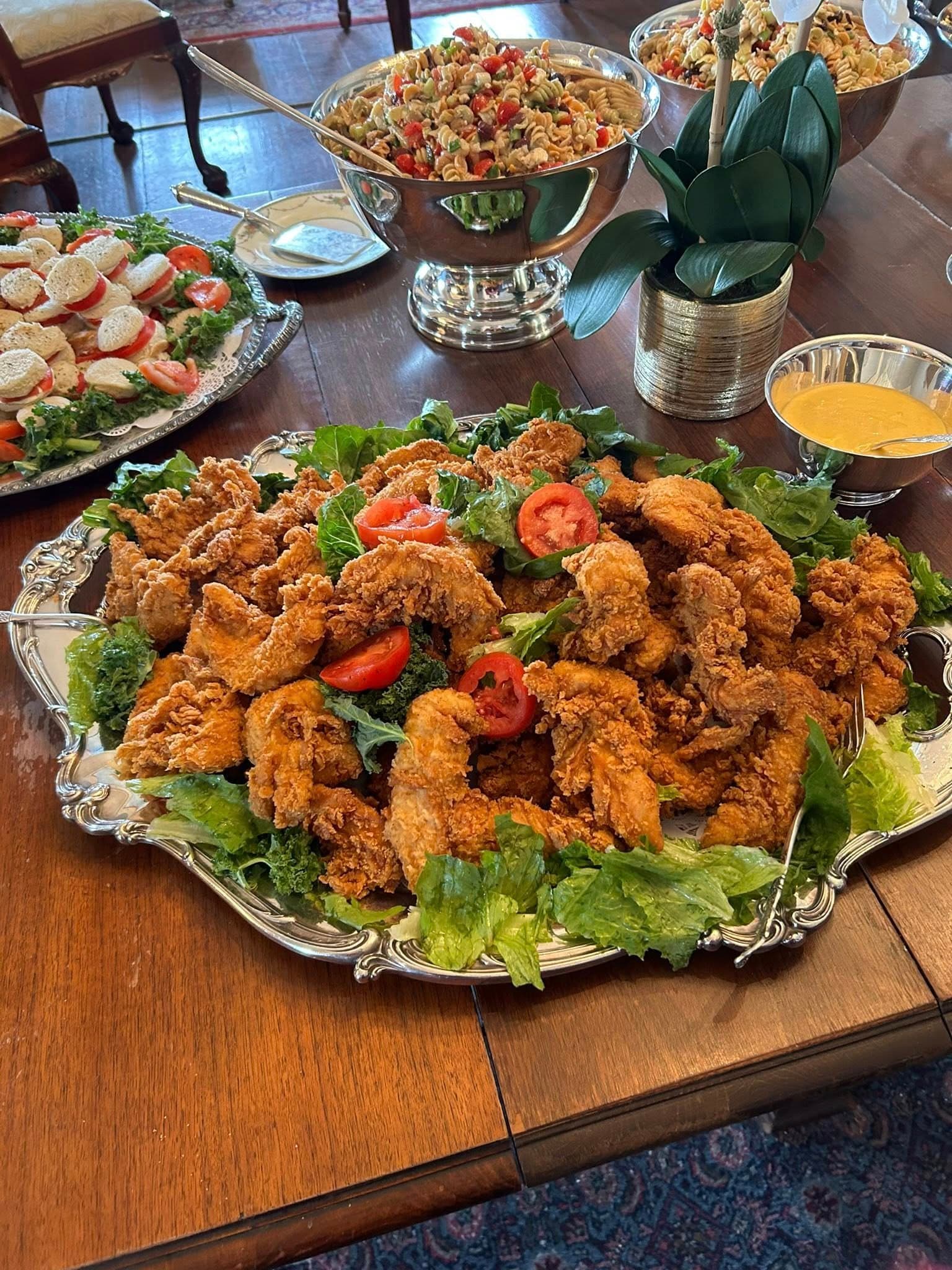 A silver platter filled with crispy fried chicken garnished with lettuce and slices of tomato, surrounded by bowls of pasta salad and salad.