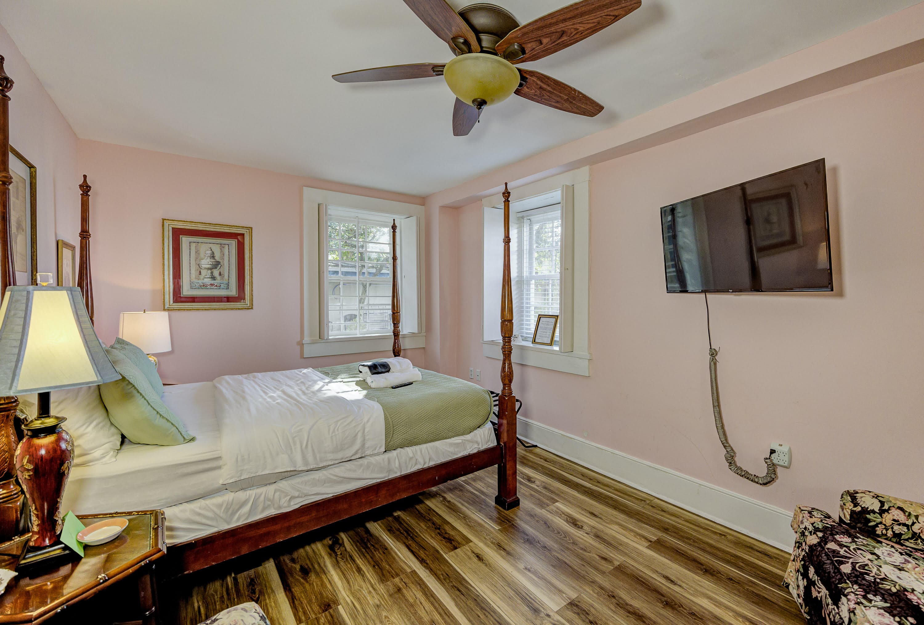 A cozy bedroom with a four-poster bed, a television mounted on the wall, and soft pink walls.