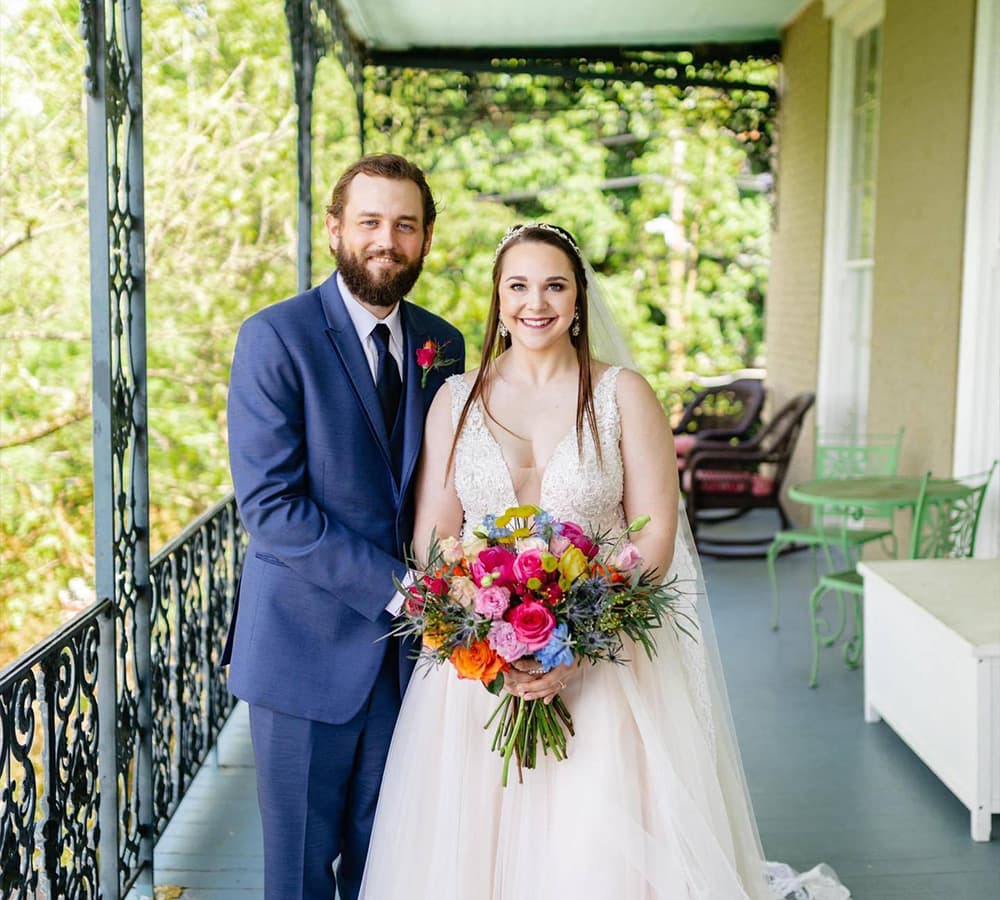A bride and groom pose together on a balcony, with the bride holding a vibrant bouquet of flowers.