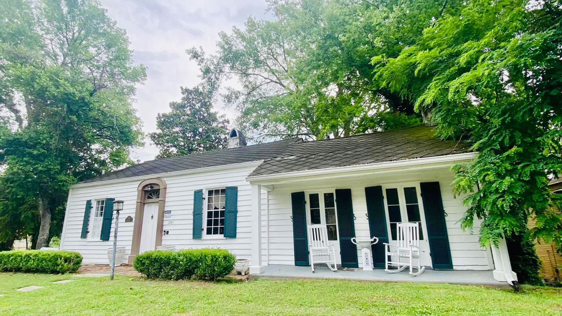 A white house with green shutters and rocking chairs on the porch, surrounded by lush greenery.