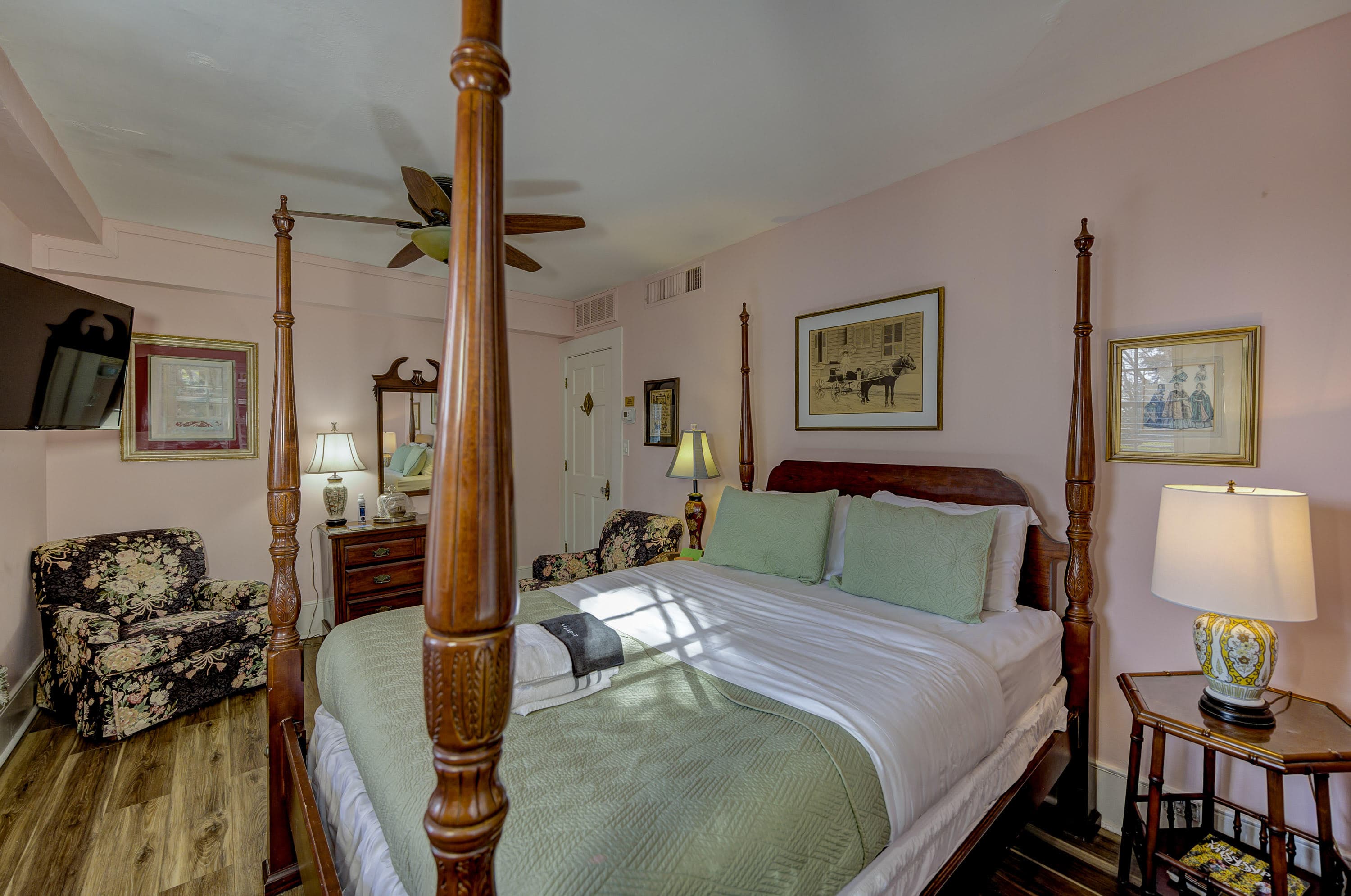 A cozy bedroom featuring a four-poster bed, floral chairs, and soft lighting against a pink wall.