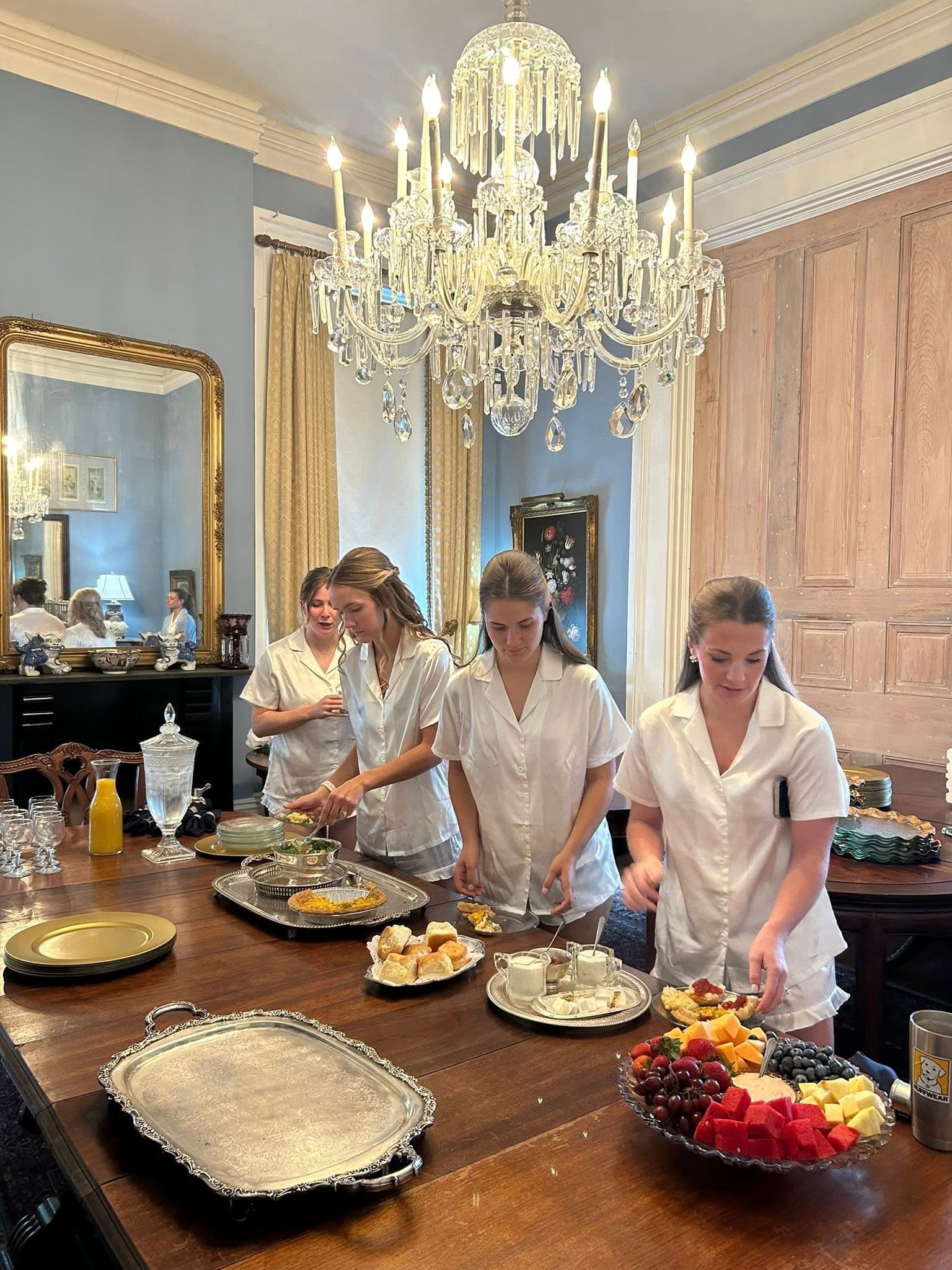 Three women in white shirts prepare food at a dining table under a chandelier.