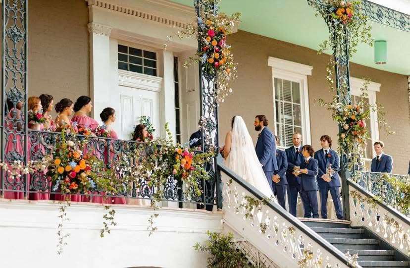 A bride and groom stand on a staircase during their wedding ceremony, surrounded by a decorated balcony and attending guests.
