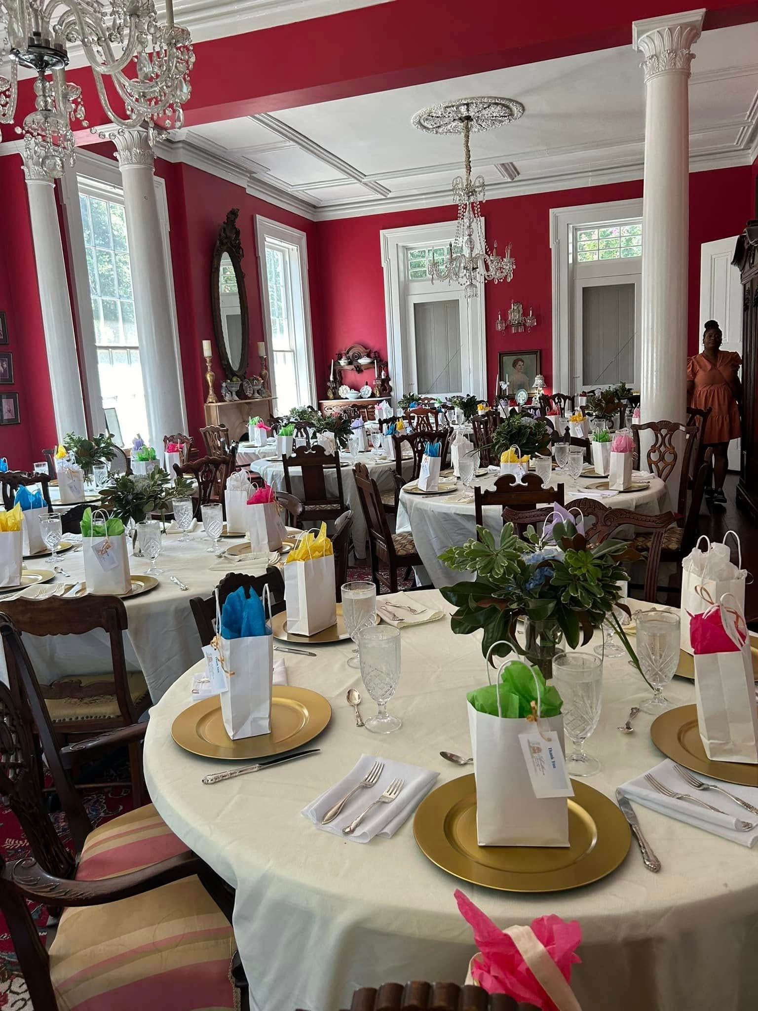 A beautifully decorated dining room with several tables set for a gathering, featuring colorful gift bags and floral centerpieces against a rich red backdrop.