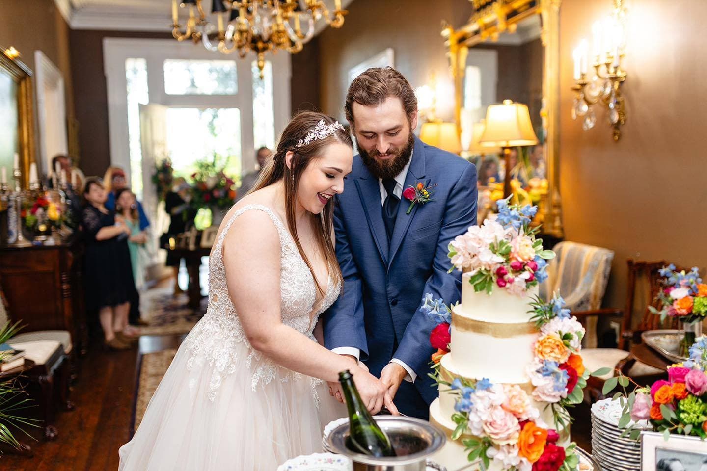 A bride and groom are smiling as they cut their wedding cake together, surrounded by floral decorations and guests.