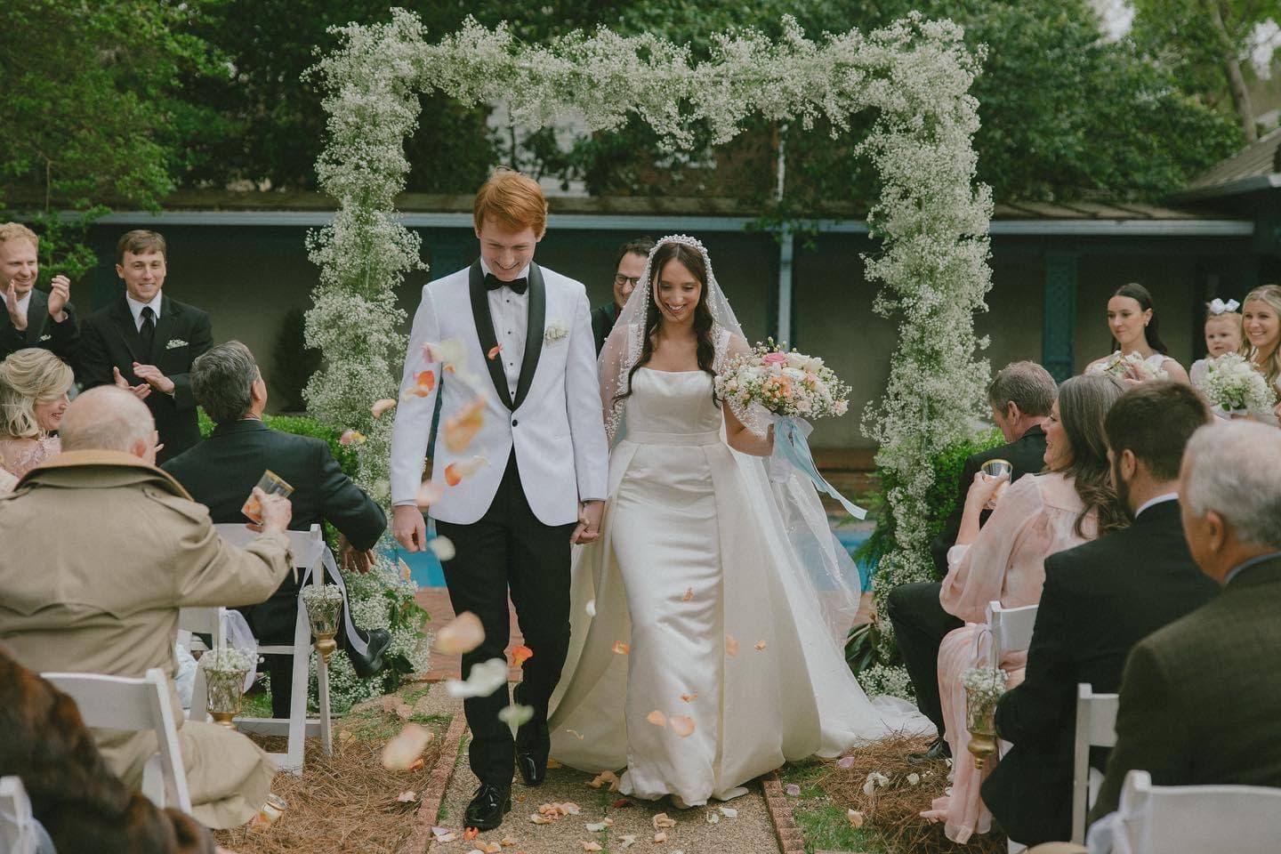 A bride and groom walk hand in hand through a flower petal-strewn aisle, surrounded by smiling guests.