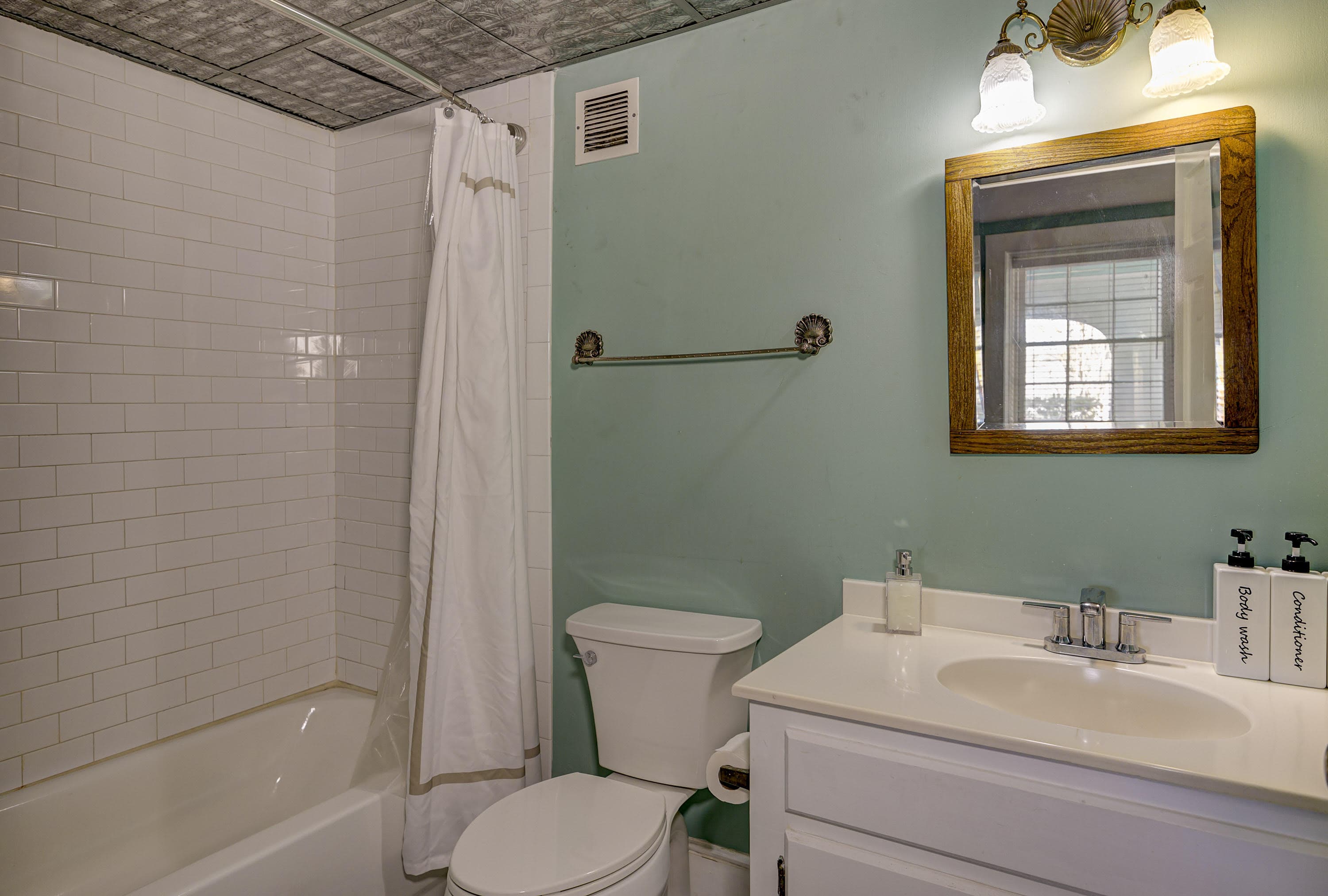 A small, modern bathroom featuring a bathtub with a shower, white tiles, a toilet, and a vanity with a mirror and toiletries.