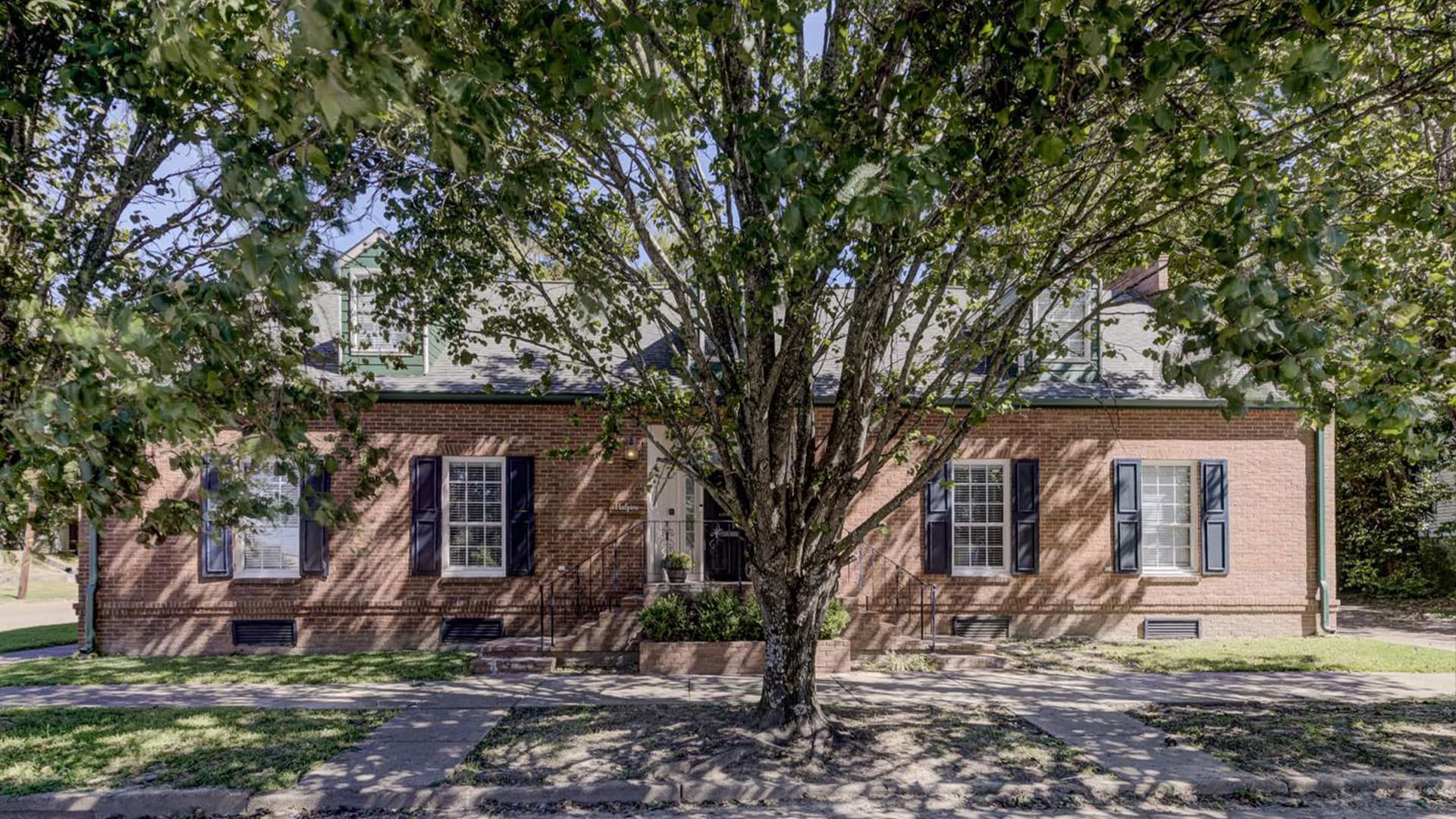A brick house with a large tree in front, featuring multiple windows and a welcoming entrance.