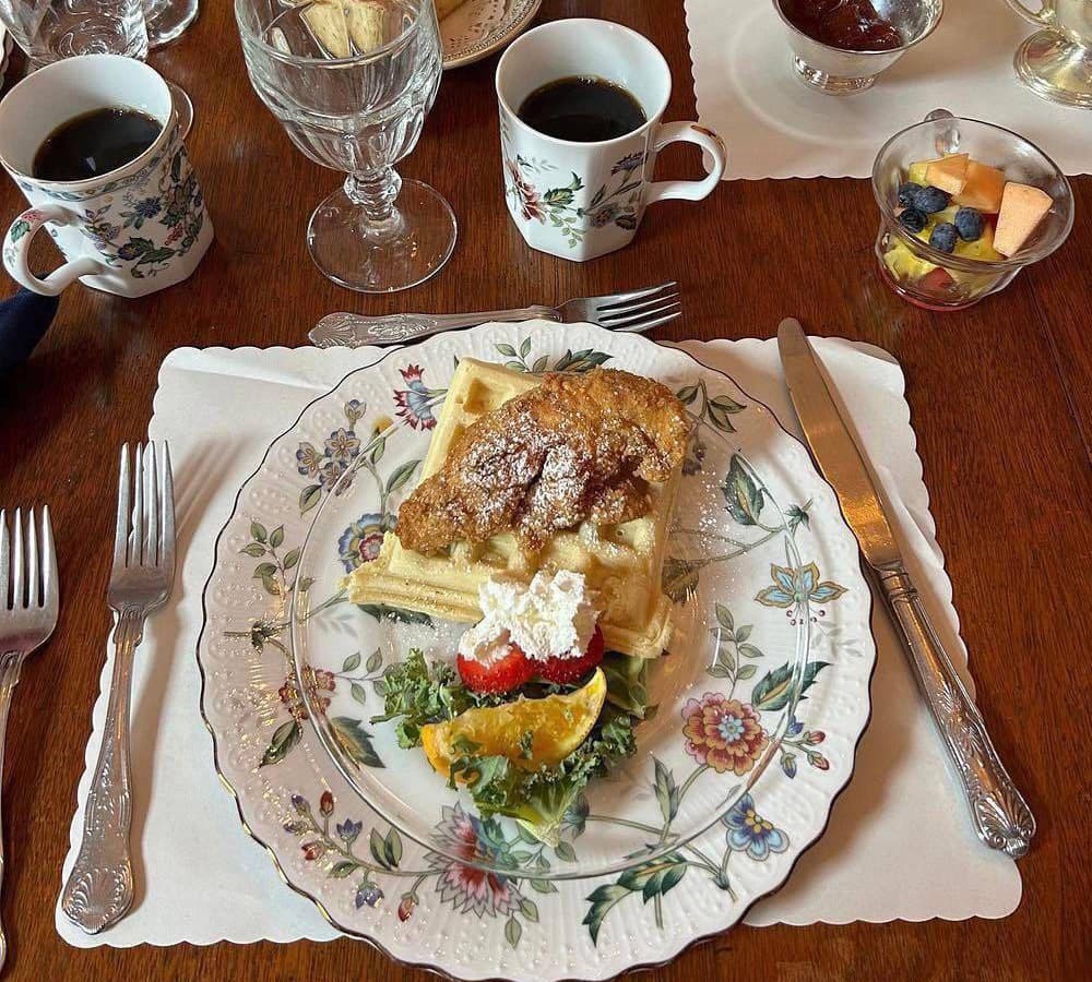 A beautifully arranged plate of waffles topped with a croissant, accompanied by fresh fruit and whipped cream, on an elegant floral table setting.