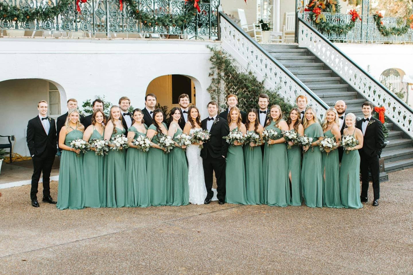 A wedding party poses together outdoors, featuring bridesmaids in green dresses and the bride and groom in formal attire.