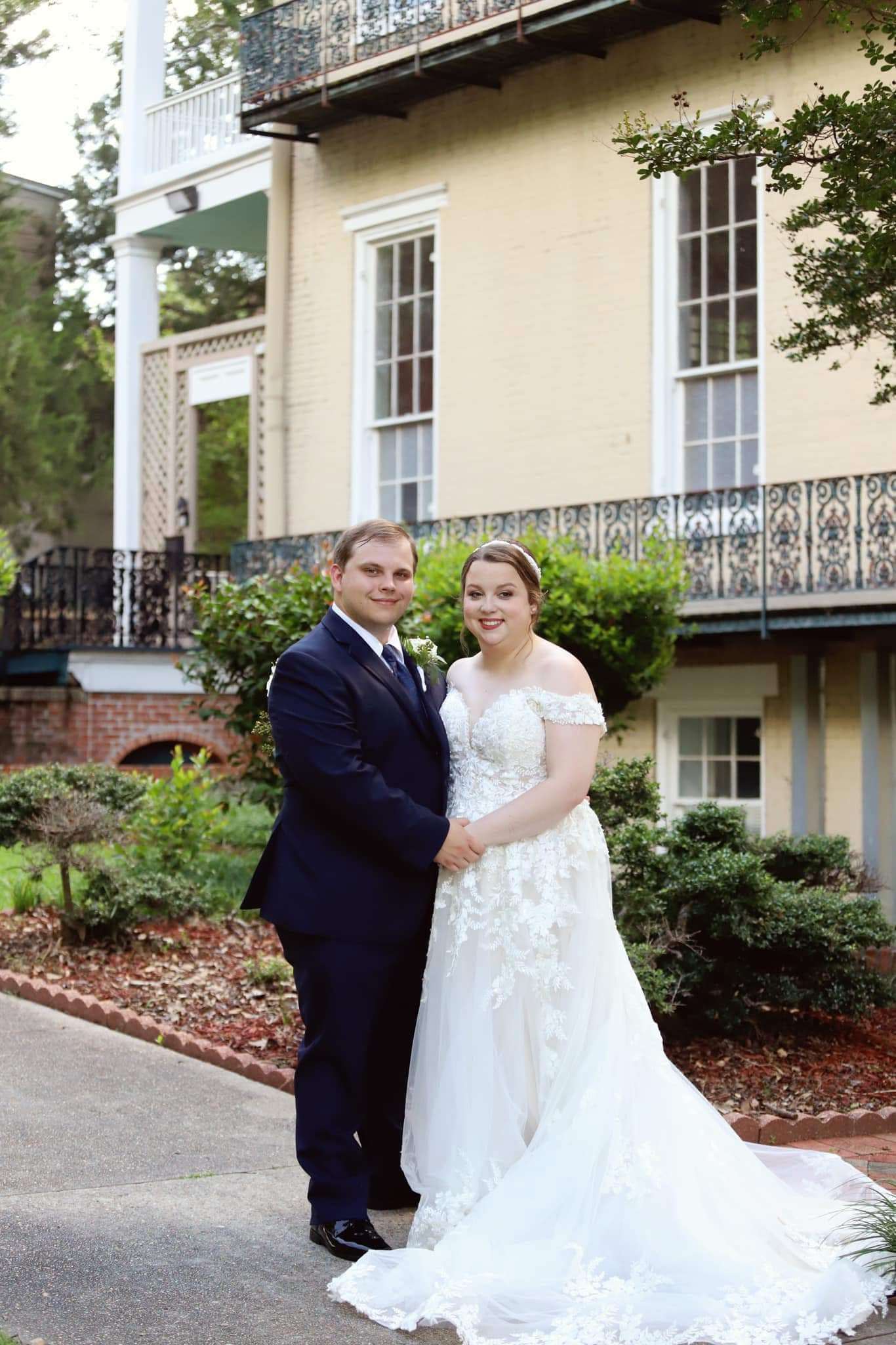 A smiling couple poses together outside a historic building, with lush greenery surrounding them.