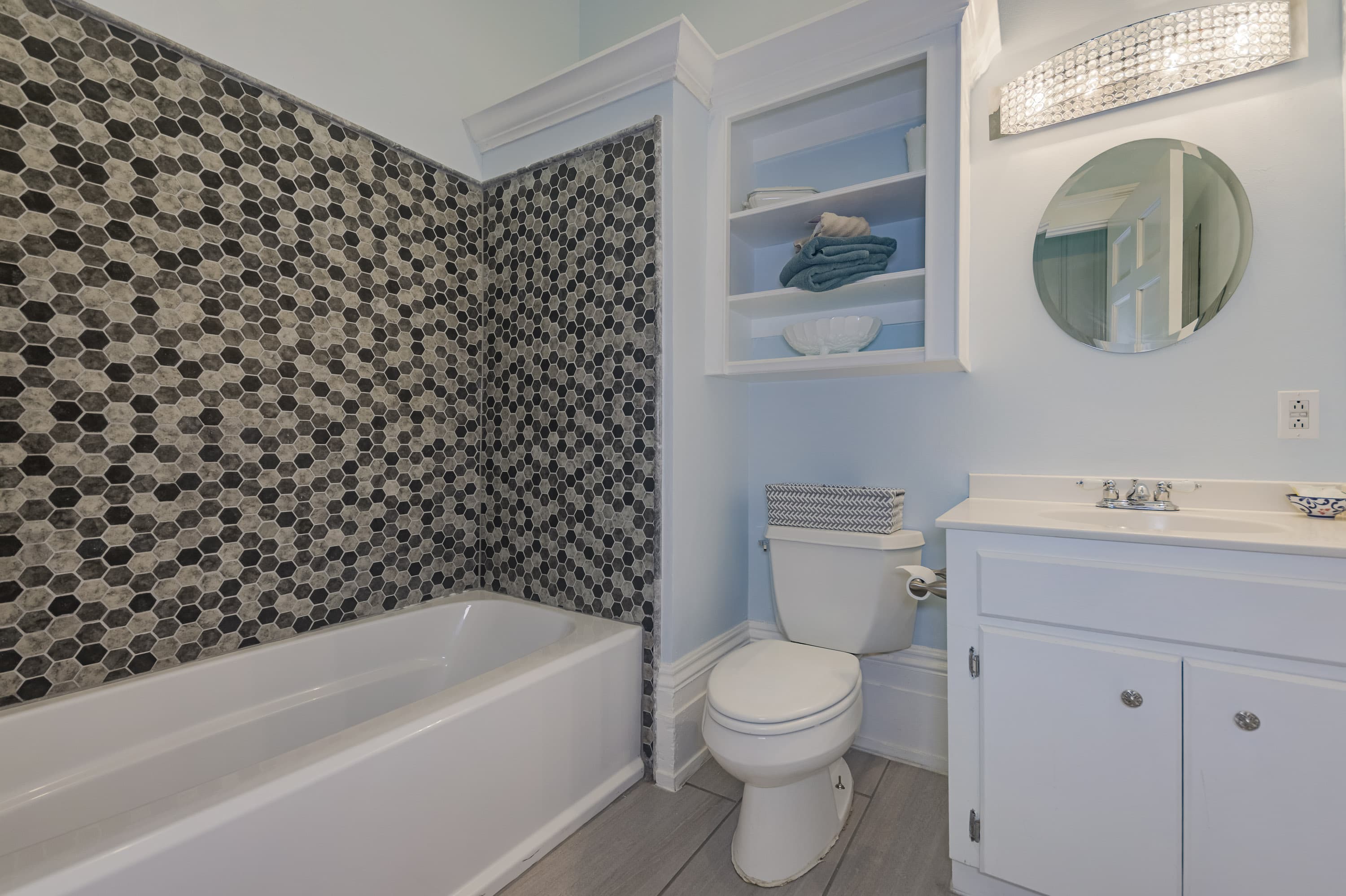 blue walls, white tub, brown surround around tub, white vanity, long stainless light, oval mirror, shelves over toilet