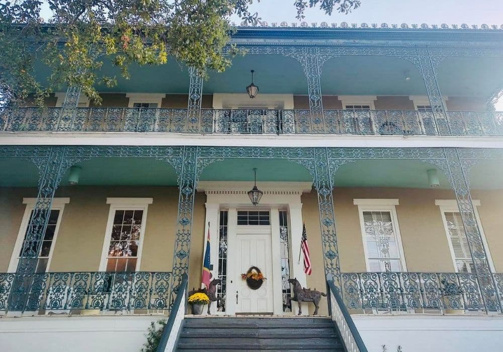 Historic two-story house with intricate blue ironwork and a white front door, decorated with a wreath, surrounded by flags and greenery.